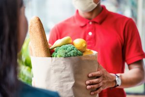 Asian deliver man wearing face mask in red uniform handling bag of food, fruit, vegetable give to female costumer in front of the house. Postman and express grocery delivery service during covid19